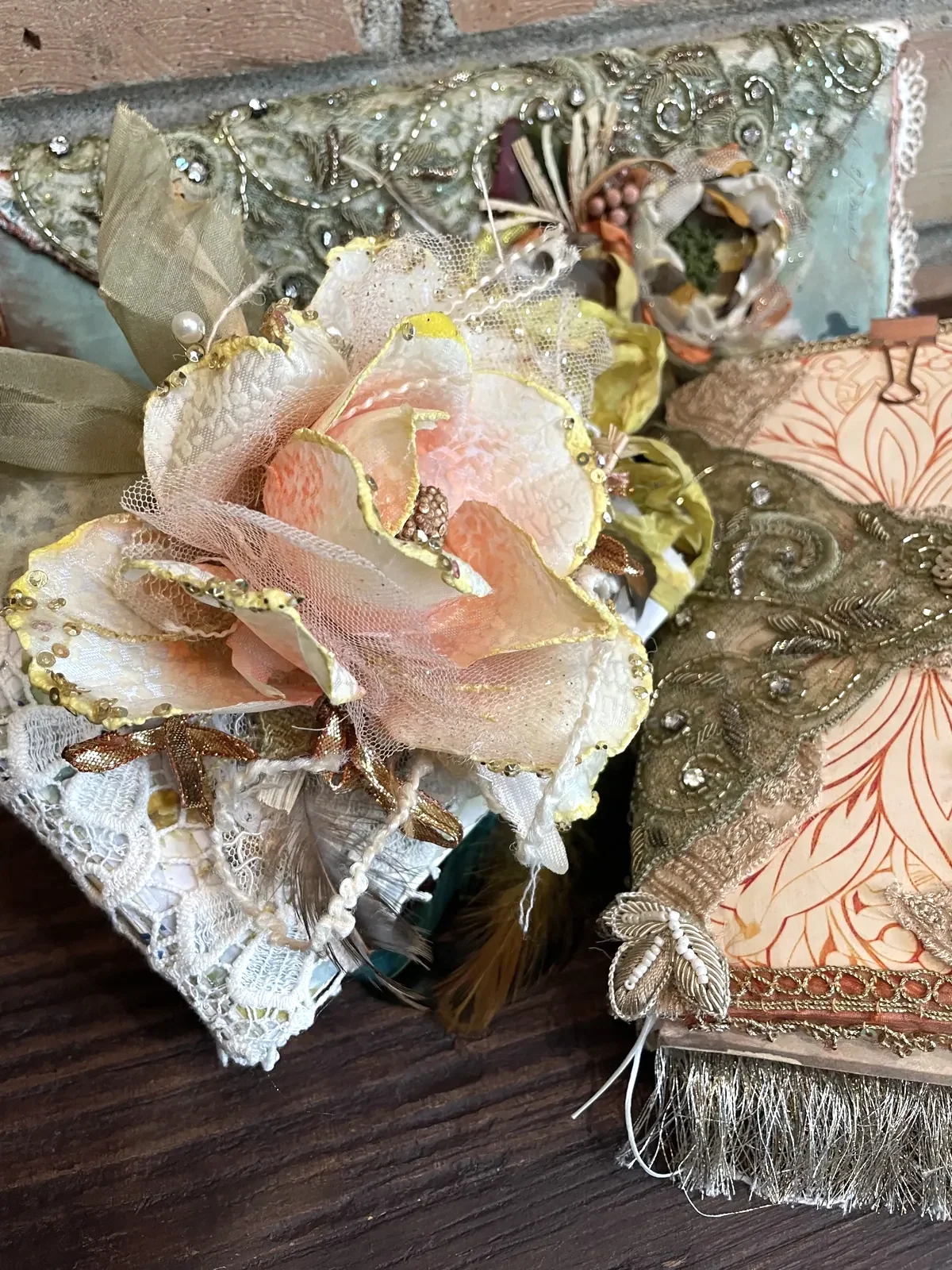 A close view of three embellished artist journals with handmade flowers, lace, embroidery and metallic trim